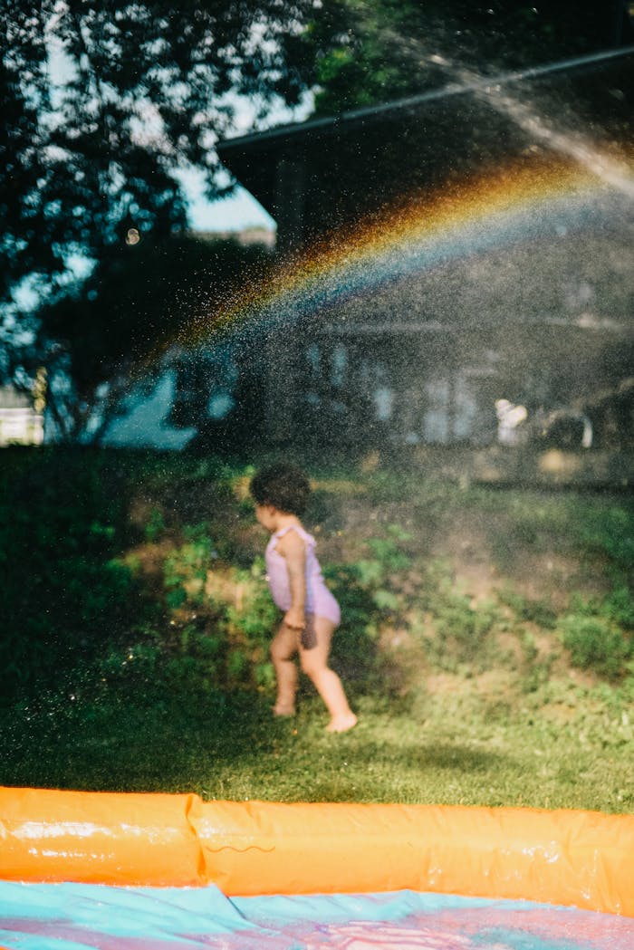 Young child playing in the backyard with a sprinkler creating a rainbow effect.